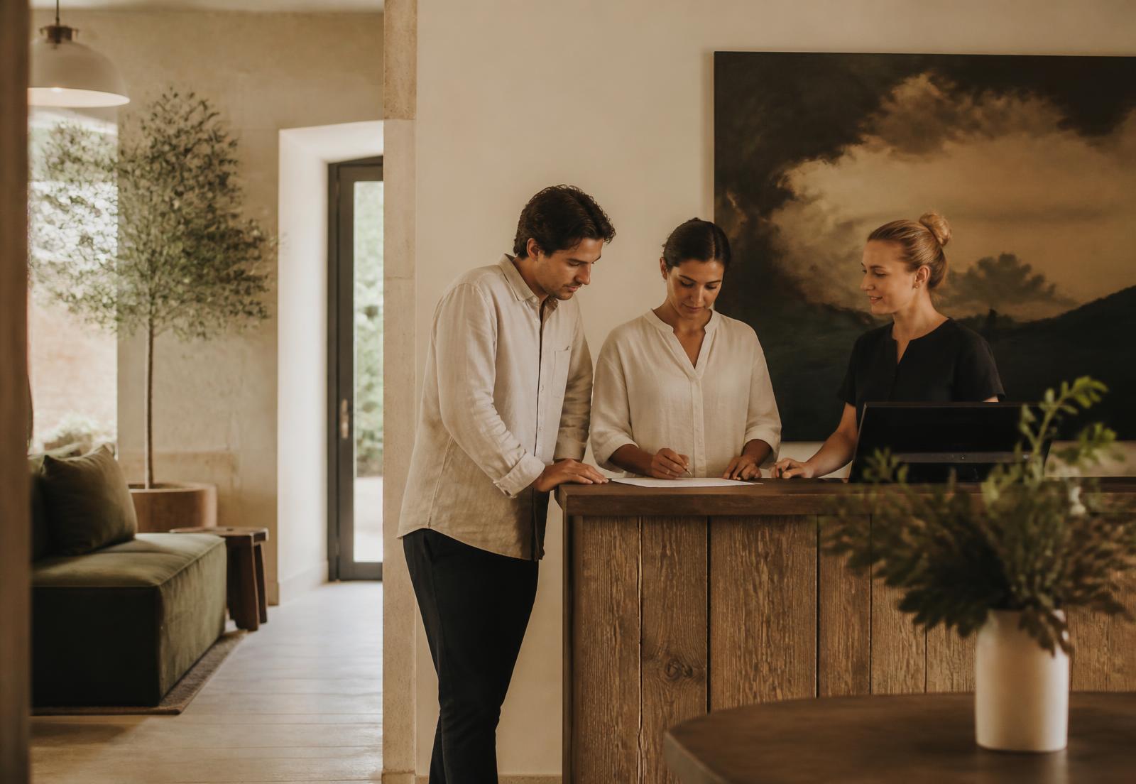 A small hotel team coordinating a guest arrival at a timber reception desk in a warm, light-filled lobby