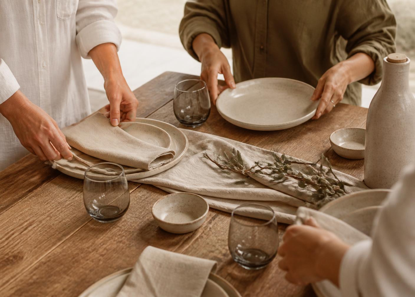 Several pairs of hands setting a timber table together with linen, ceramics and glassware